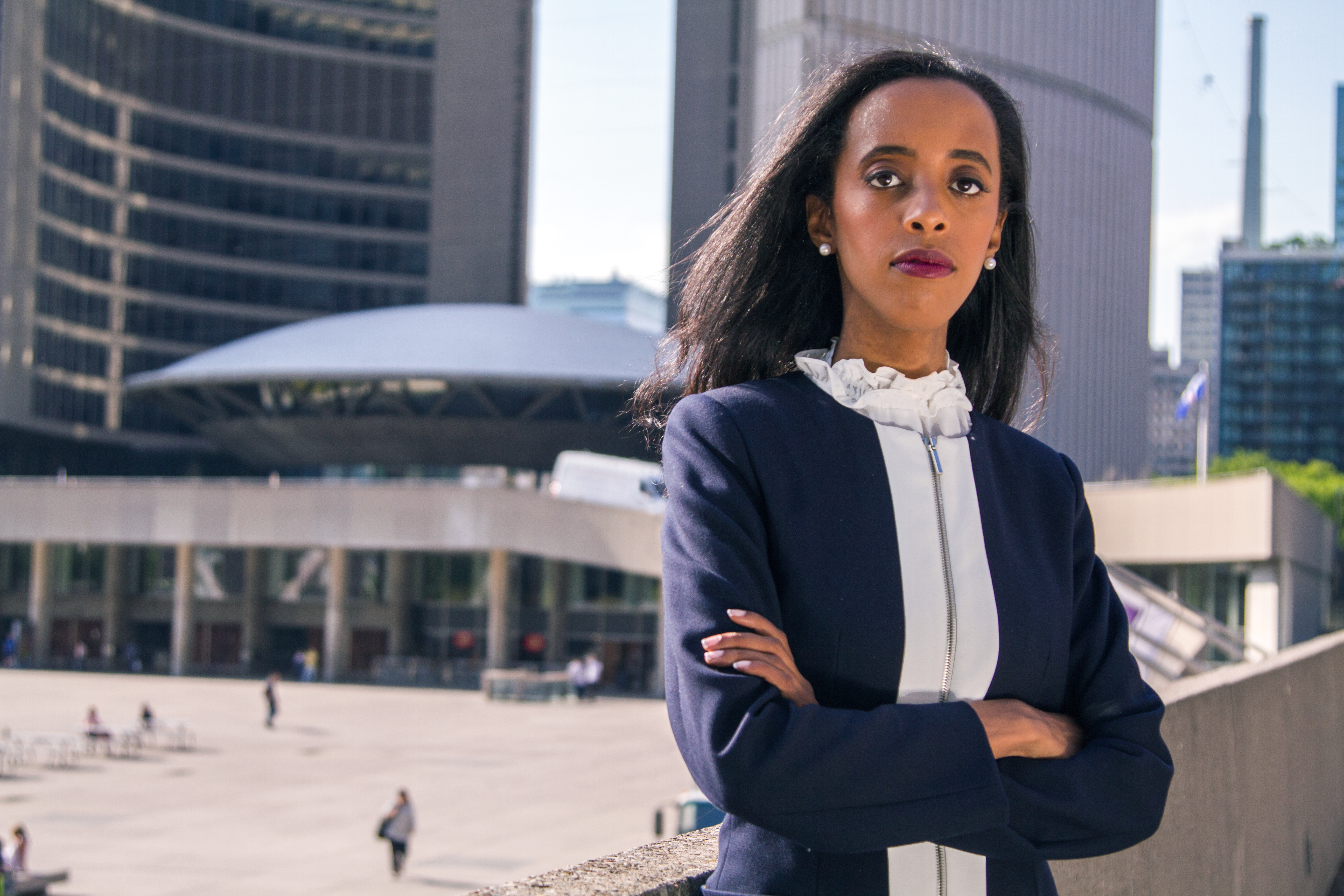 Saron Gebresellassi City of Toronto 2018 mayoral candidate stands in front of City Hall with her arms crossed whilie looking with serious expression into camera. The background of the City Hall building is slightly blurred with some citizens walking about.